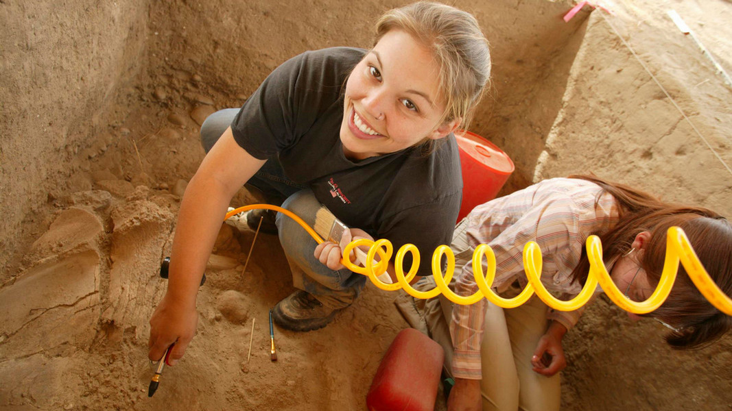 A student smiles from ear to ear as she is crouched next to another student within a ditch,