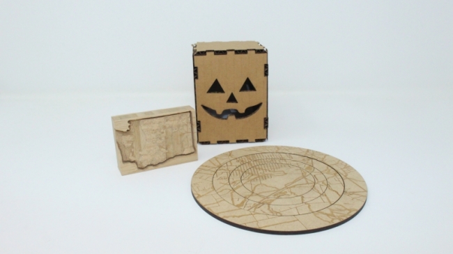 various laser cutting and etching examples including a pumpkin box made of cardboard, a circular puzzle with the map of Kittitas County etched on it and block of wood with the state of Washington carved into it
