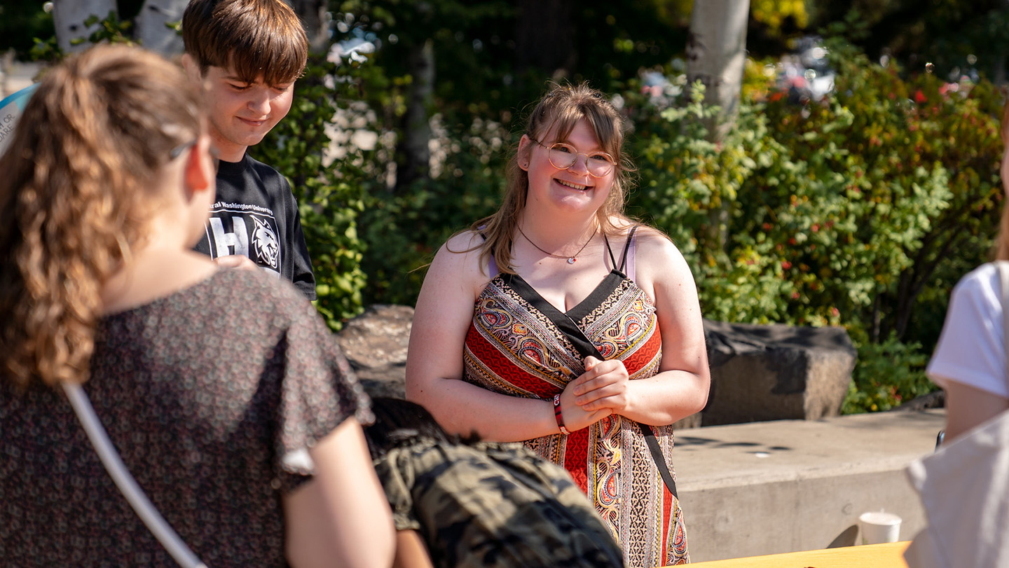 A girl is smiling and laughing with friends outside. There are bushes behind them and she is wearing glasses.