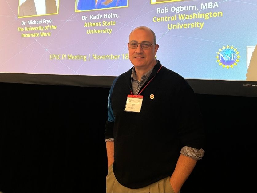 BCS Executive Director Rob Ogburn poses in front of a projection screen, equipped with a name tag. The projection screen lists the members of the EPIIC PI convention's community engagement panel, including Ogburn.