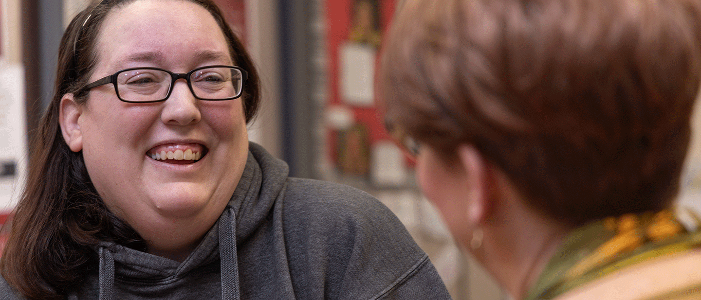 A smiling adult learner wearing glasses and a university hoodie talks with an advisor in a welcoming campus setting, suggesting personalized academic support and guidance.