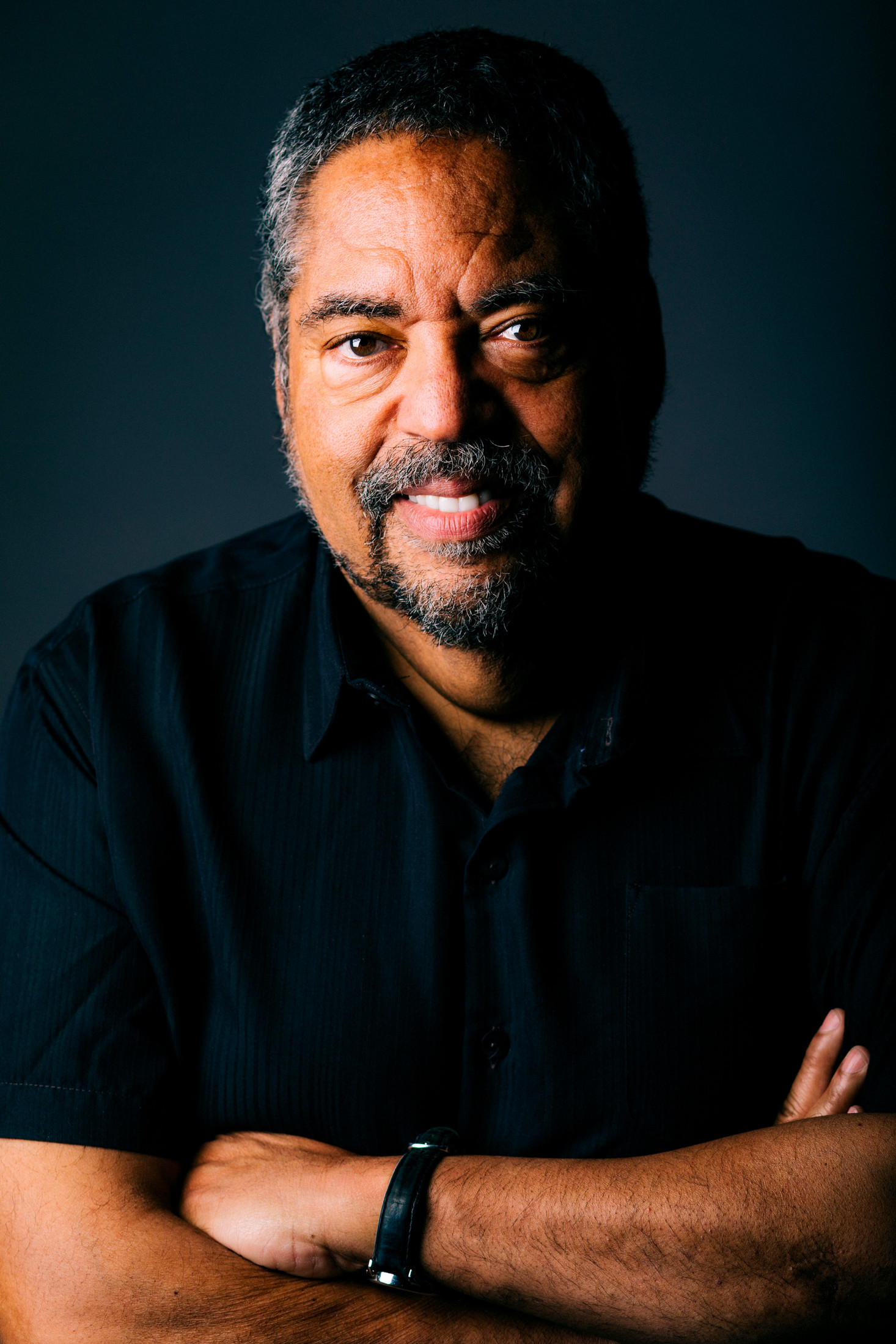 Portrait of David Haynes wearing black, against black background with his arms folded and part of his face obscured as he smiles at the camera