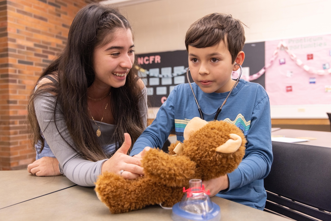 Student holding a teddy bear that is being looked at by a young child with a stethoscope on